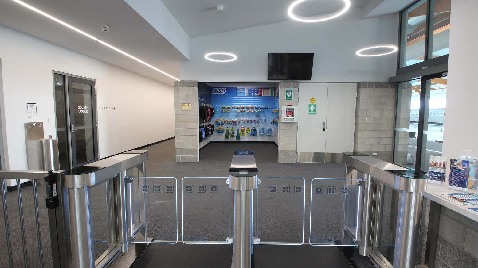 A closer look at the turnstiles, including an accessible turnstile that leads to a small retail space selling a range of aquatic gear. On the upper wall is a television. There is a white double door with a green first aid sign on the right of the retail space. There is a water fountain station accessible on the left corridor that leads to Changerooms. Right before the water fountain stain is the tinted door of SQUASH Courts.