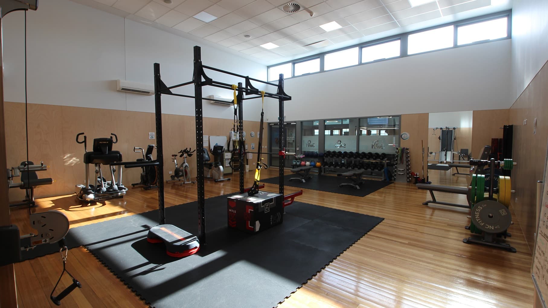 The timber floor inside the gym of SWIRL is well polished. The gym has a chic-vibe design with wood-like brown and white painted walls. The ceiling is composed of a small rectangular design that is painted in white. Found inside the gym are kettlebells, black adjustable professional gym benches, treadmills, bicycles, and a multi-functional squat power rack. There is a large mirror at the back, and some floor areas are covered by large black mats.