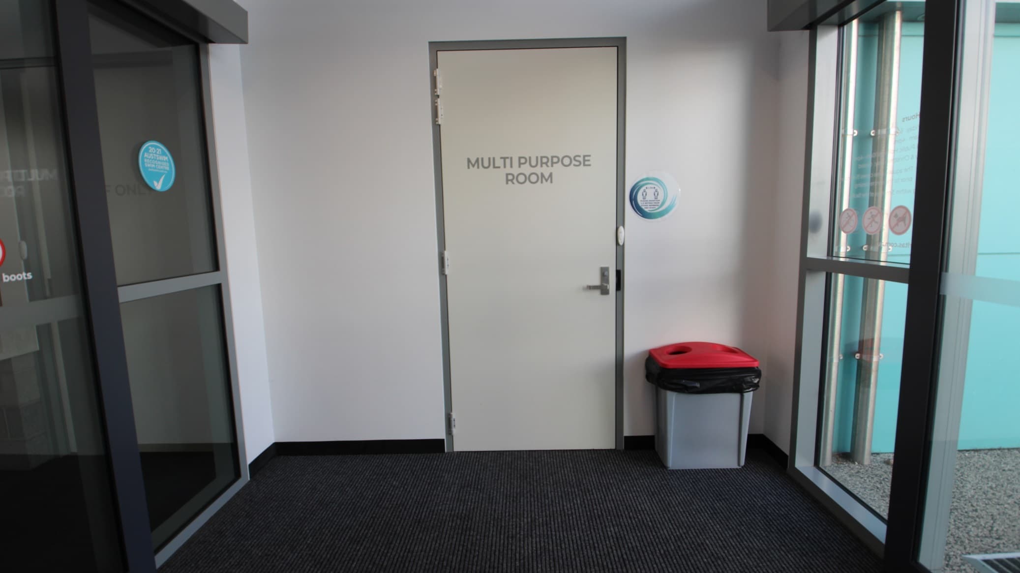 The white door of the Multipurpose room at SWIRL can be found on the right after passing through the automatic glass double doors at the entrance. There is a rubbish bin with a black garbage bag and a red lid on the right of the multipurpose door.