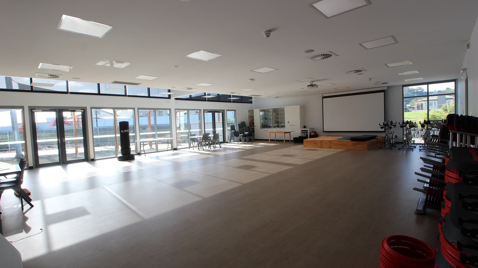 Inside the Multipurpose room is a large, open space. There is a wooden block in front of the large projector screen. On the left is a white cabinet with a stack of black chairs with steel legs. On the right are stationary bicycles for exercise. Natural light is filtered through the glass walls and windows.