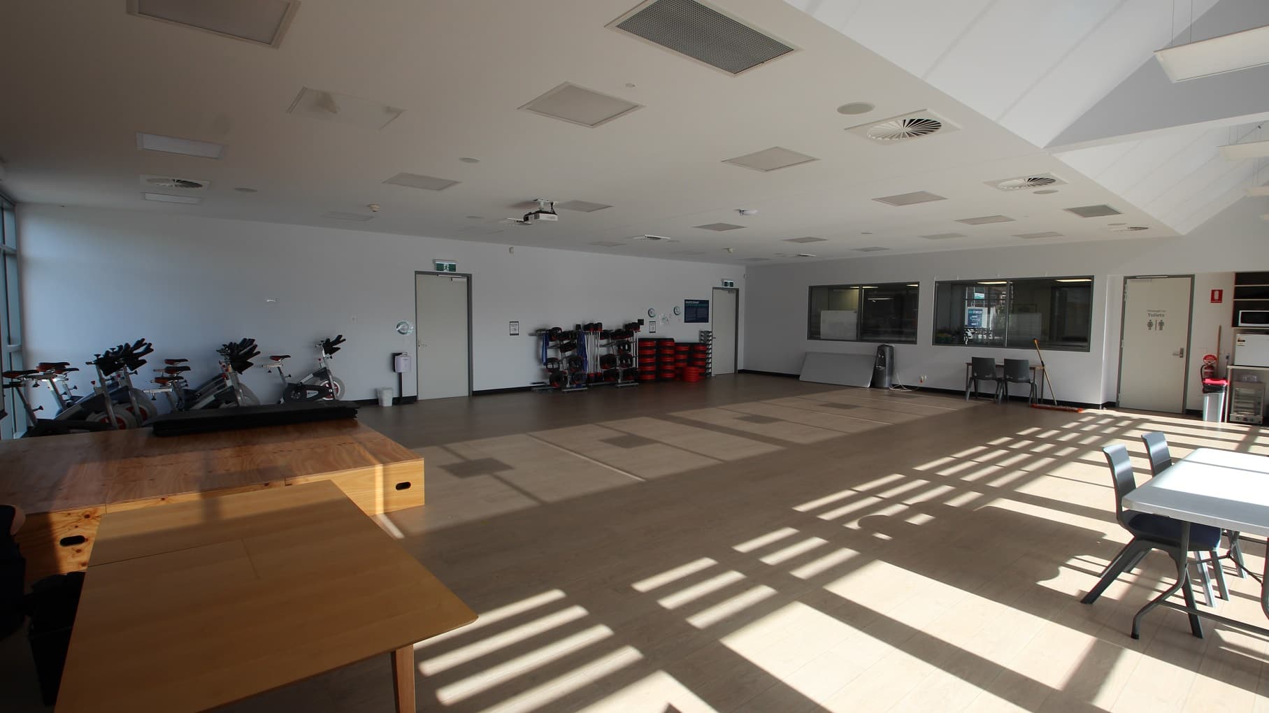 There are three doors inside the Multipurpose room. There is a wooden block in front of the large projector screen, and white tables with grey chairs are accessible in the room. On the right are stationary bicycles for exercise. Natural light is filtered through the glass walls and windows.
