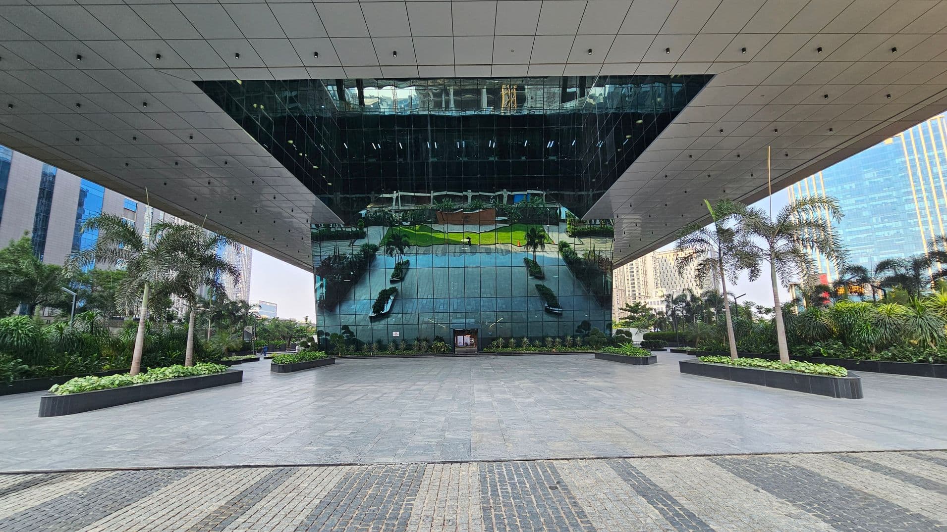 A modern architectural structure with a large reflective glass facade in the centre, mirroring the surrounding area. The foreground features a spacious tiled plaza with a patterned design. On both sides, there are rectangular planters with palm trees and lush greenery. The ceiling is composed of a grid of panels with small lights. In the background, tall buildings are visible, adding to the urban setting.