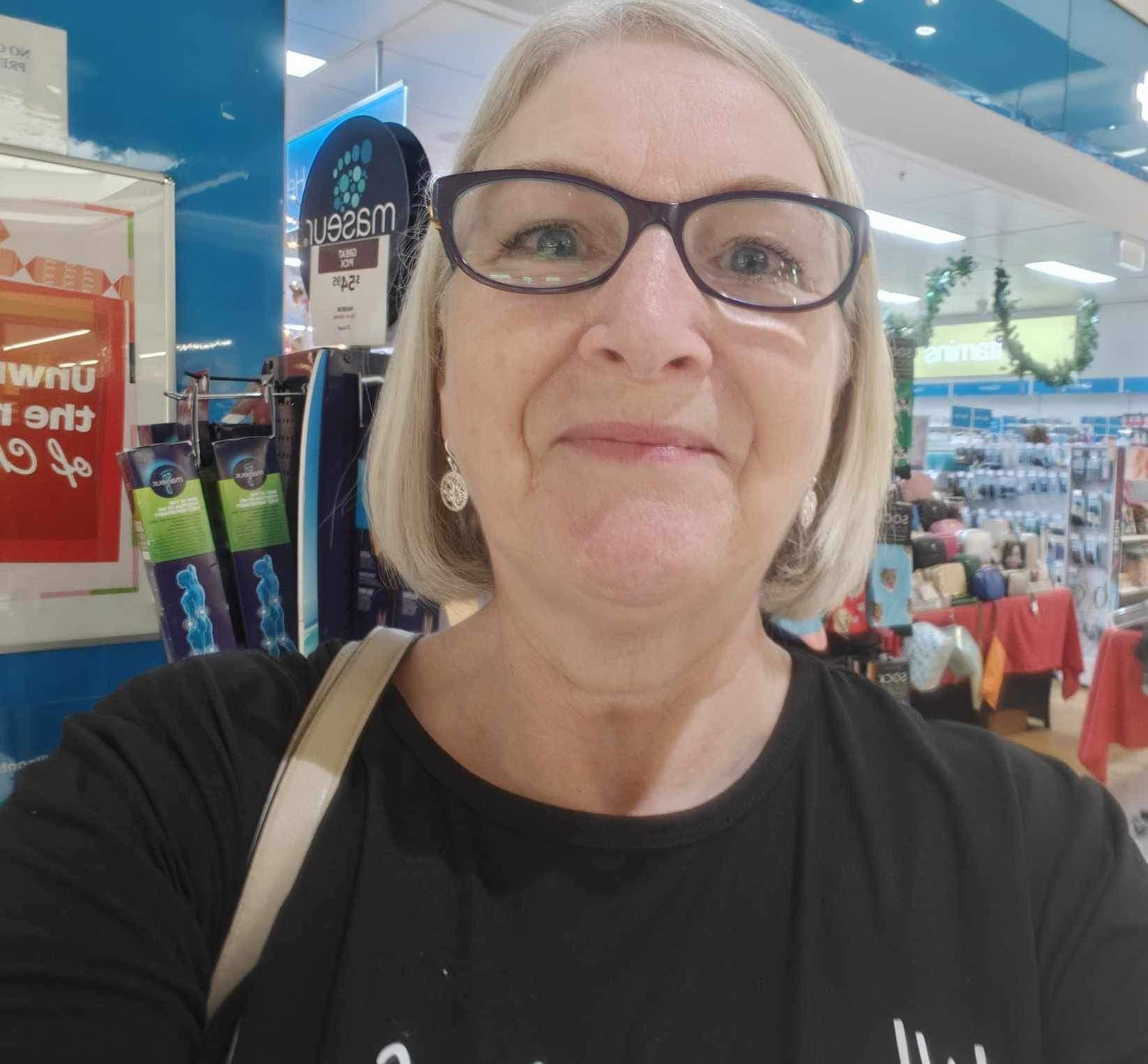 A woman with short hair and glasses stands in front of a pharmacy.