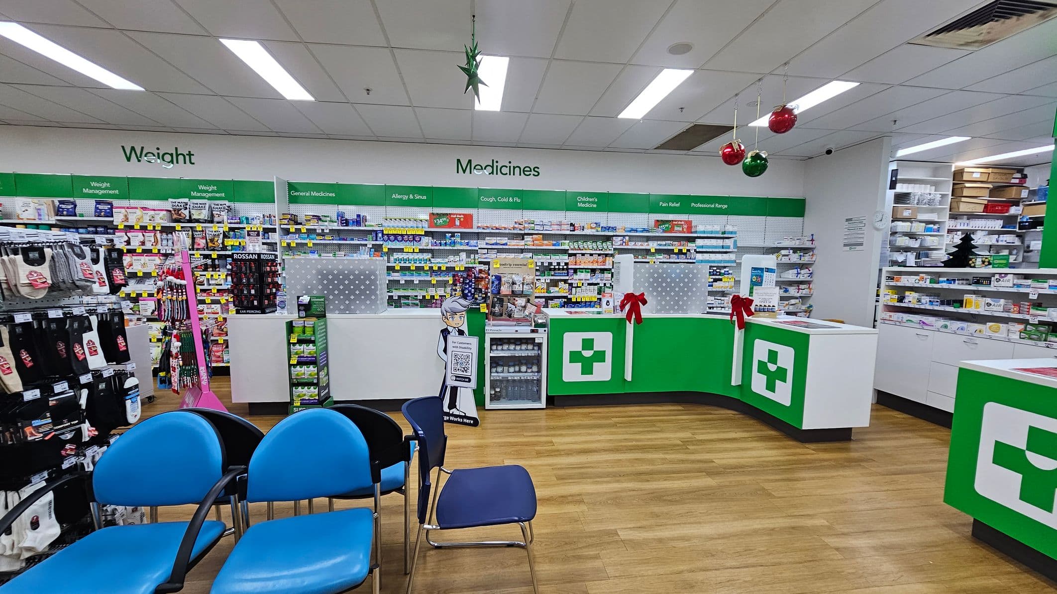 Pharmacy interior with a wooden floor and a white ceiling featuring fluorescent lights. On the left, blue chairs are arranged in a waiting area. The back wall has shelves stocked with various products under signs like "Weight" and "Medicines." The counter in the centre has a green and white design with a medical cross symbol, decorated with red ribbons. Hanging from the ceiling are festive decorations, including red and green baubles.