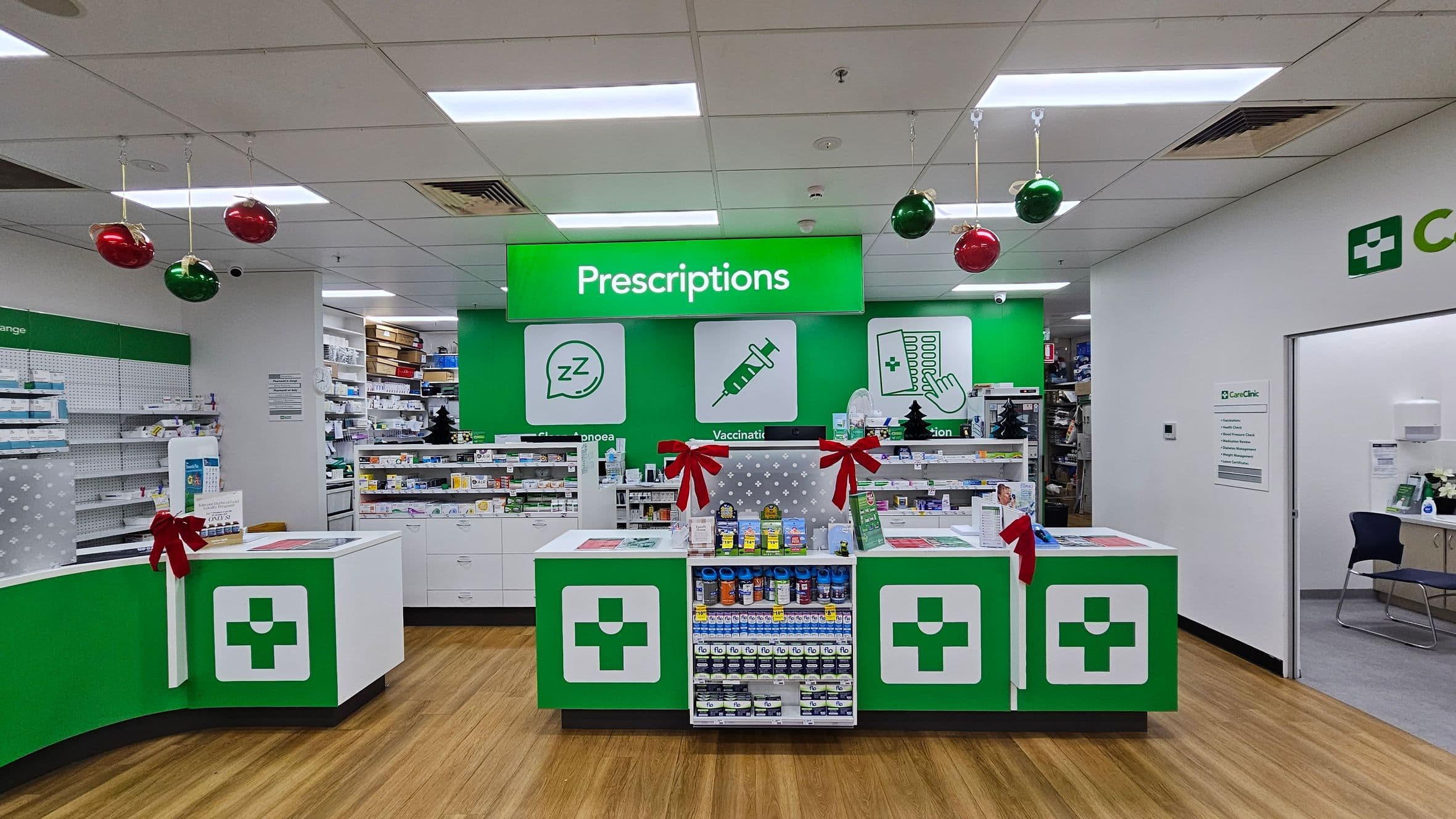 Pharmacy interior with a wooden floor and a white ceiling featuring recessed lighting. In the centre, a green sign reads "Prescriptions" above a counter with green and white cross symbols. The counter displays various products and is decorated with red bows. Shelves filled with medicines are visible in the background. Hanging from the ceiling are red and green baubles. On the right, a doorway leads to a room with a chair and a sign labelled "CareClinic".