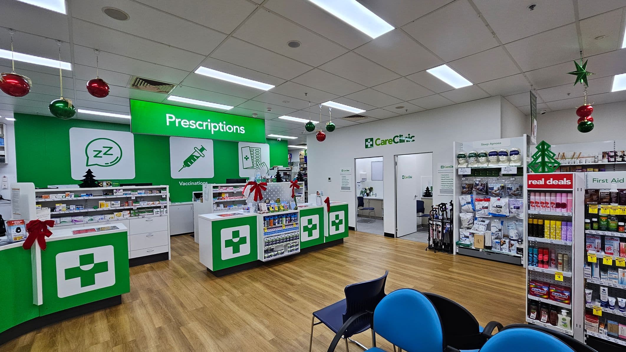 Pharmacy interior with a wooden floor and a white ceiling featuring fluorescent lights. On the left, there are green and white counters with a medical cross symbol, decorated with red bows. Behind the counters, shelves display various medications. In the centre, a large green sign reads "Prescriptions." On the right, shelves labelled "real deals" and "First Aid" hold various products. Hanging from the ceiling are red and green baubles and a star. Blue and black chairs are in the foreground.