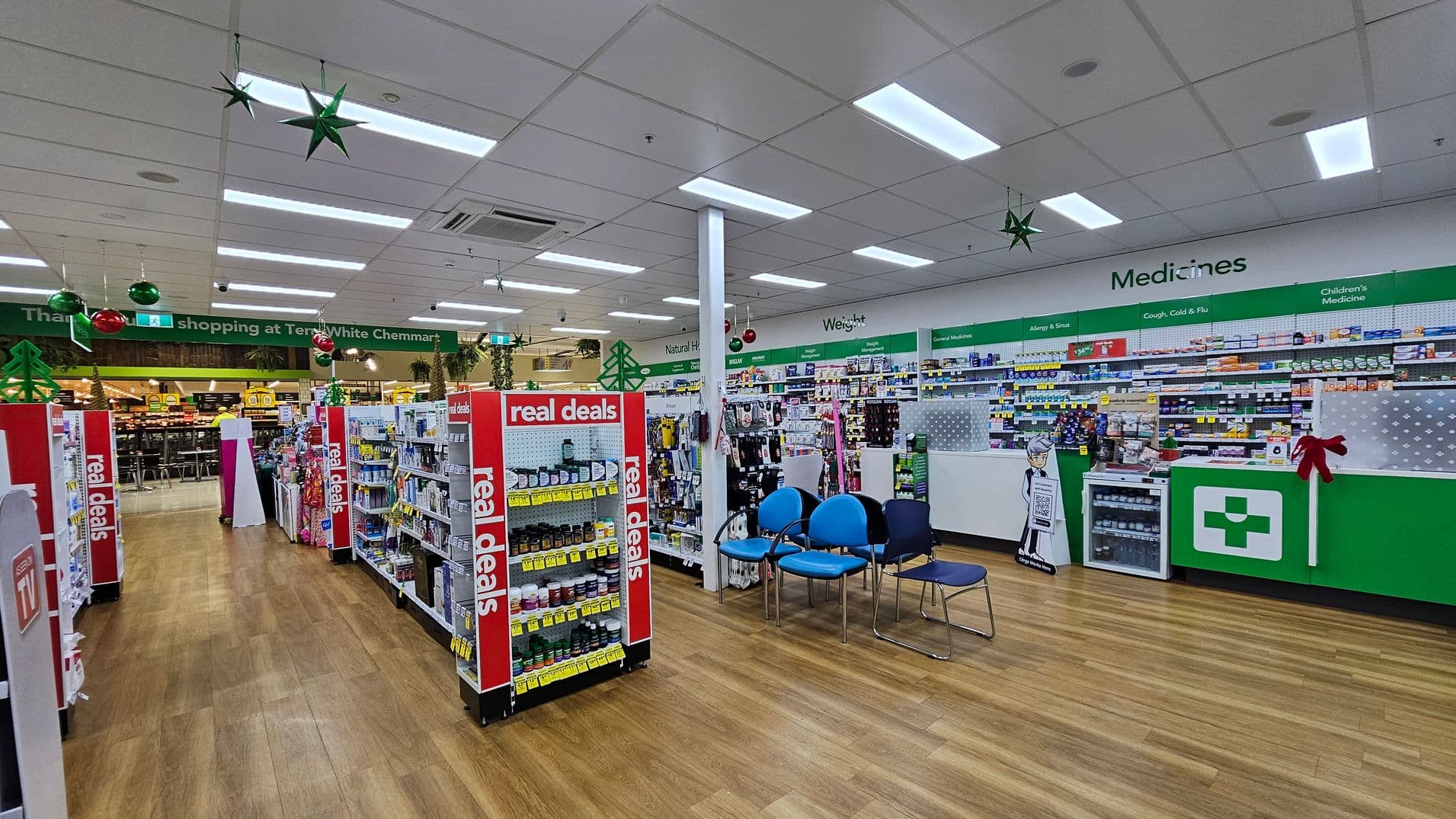 Pharmacy interior with wooden flooring and a white ceiling. On the left, shelves display products with "real deals" signs. In the centre, a seating area with blue chairs. On the right, a green and white counter with a pharmacy cross symbol, and shelves labelled "Medicines" with various health products. Hanging decorations include green stars and red baubles. In the background, a sign reads "Thank you for shopping at TerryWhite Chemmart."