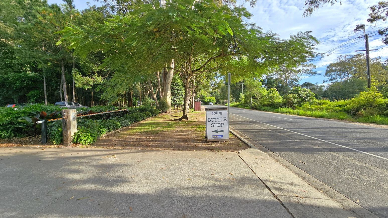 A road stretches into the distance on the right, bordered by lush greenery and trees. A sign on the pavement reads "Doonan Bottle Shop" with an arrow pointing left. On the left, a path runs parallel to the road, lined with dense foliage and a wooden post with a barrier. A vehicle is partially visible among the trees. The area is shaded by large trees, and the ground is covered with fallen leaves.