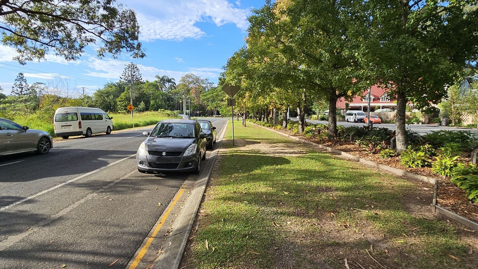 A street scene with a black car parked on the left side of the road. To the right, there's a grassy verge with trees lining the pavement. In the background, a white van and a silver car are driving on the road. Further back, a red-roofed building is partially visible behind more trees and shrubs. The road is bordered by a yellow line, and the area is well-shaded by the trees.