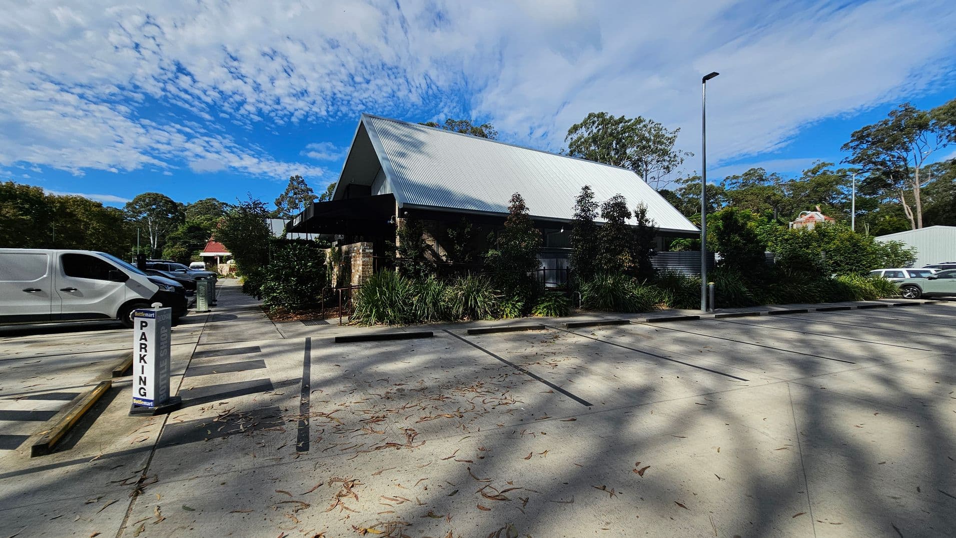A parking area with a sign marked "PARKING" on the left. Several vehicles, including a white van, are parked. In the centre, a building with a steep, corrugated metal roof is surrounded by greenery. The right side shows more parking spaces and a tall streetlamp. The ground is concrete with scattered leaves and shadows from nearby trees.