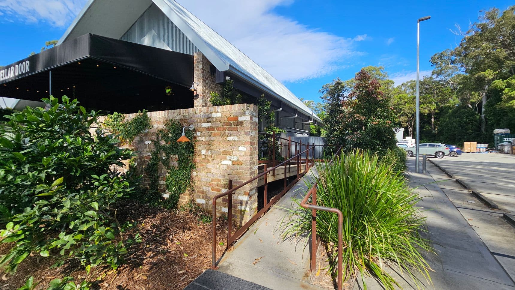 A building with a sloped metal roof and a brick exterior is on the left. A black awning with the words "CELLAR DOOR" extends from the building. A ramp with metal railings leads up to the entrance. Greenery, including bushes and climbing plants, surrounds the area. On the right, a paved area with parked cars and a tall streetlamp is visible. Trees and foliage are in the background.