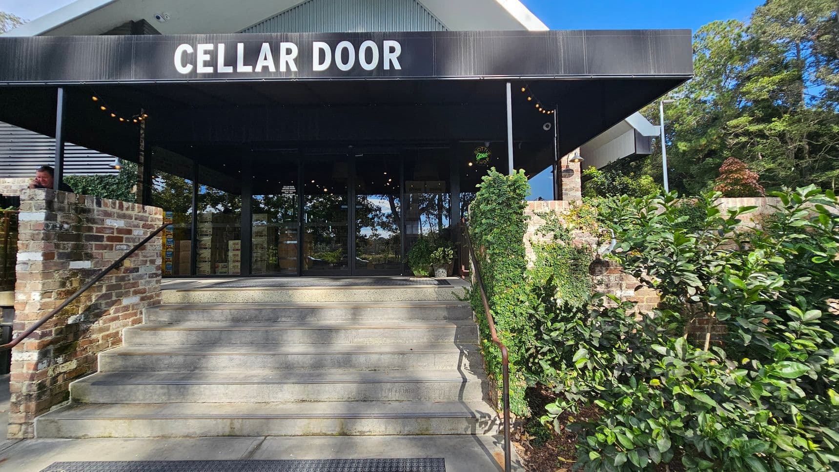 A building entrance with a sign reading "CELLAR DOOR" above. The entrance is accessed by a set of concrete steps flanked by brick walls. On the right, green foliage and vines cover part of the wall. The left side shows a person leaning on the brick wall. The background features glass doors reflecting trees and greenery. The ceiling of the entrance is dark, with string lights visible.