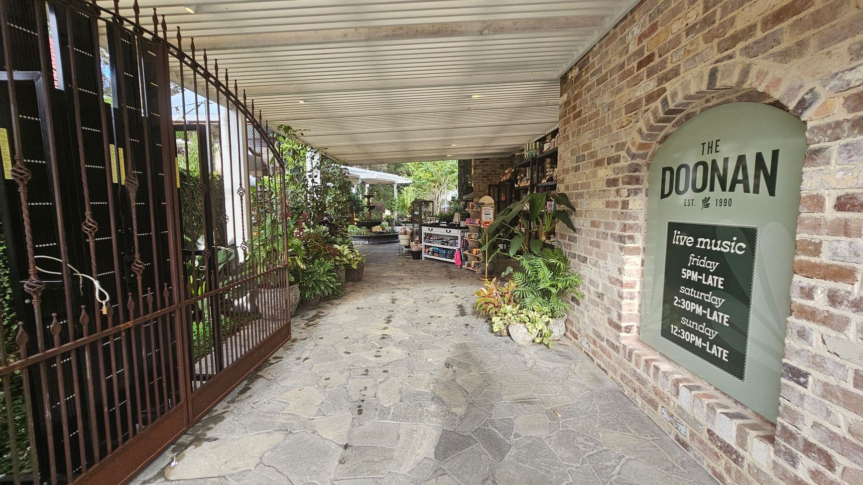 A covered walkway with a stone floor leads to a garden area. On the left, there's a wrought iron gate with decorative elements. On the right, a brick wall features a sign for "The Doonan" with live music timings. Various plants and greenery are placed along the path, and shelves with items are visible. The ceiling is wooden with recessed lighting. In the background, more plants and a seating area can be seen.