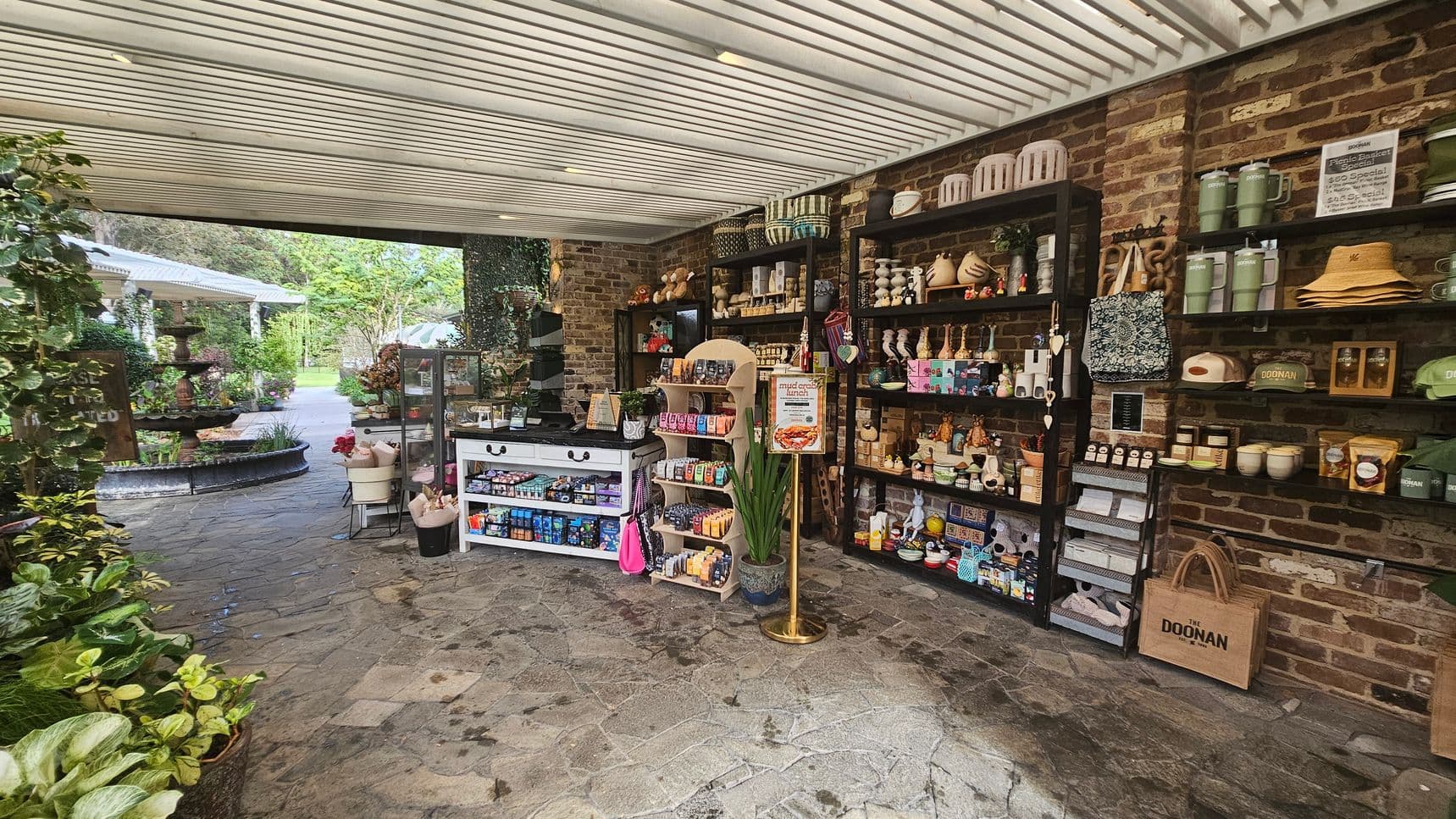 A small shop with a rustic brick wall on the right, featuring shelves filled with various items like mugs, hats, and candles. In the centre, a white display table and a wooden rack hold snacks and small goods. The floor is stone-tiled. On the left, lush green plants and a path lead outside to a garden area with a fountain and more greenery. The ceiling is slatted, allowing natural light to filter through.