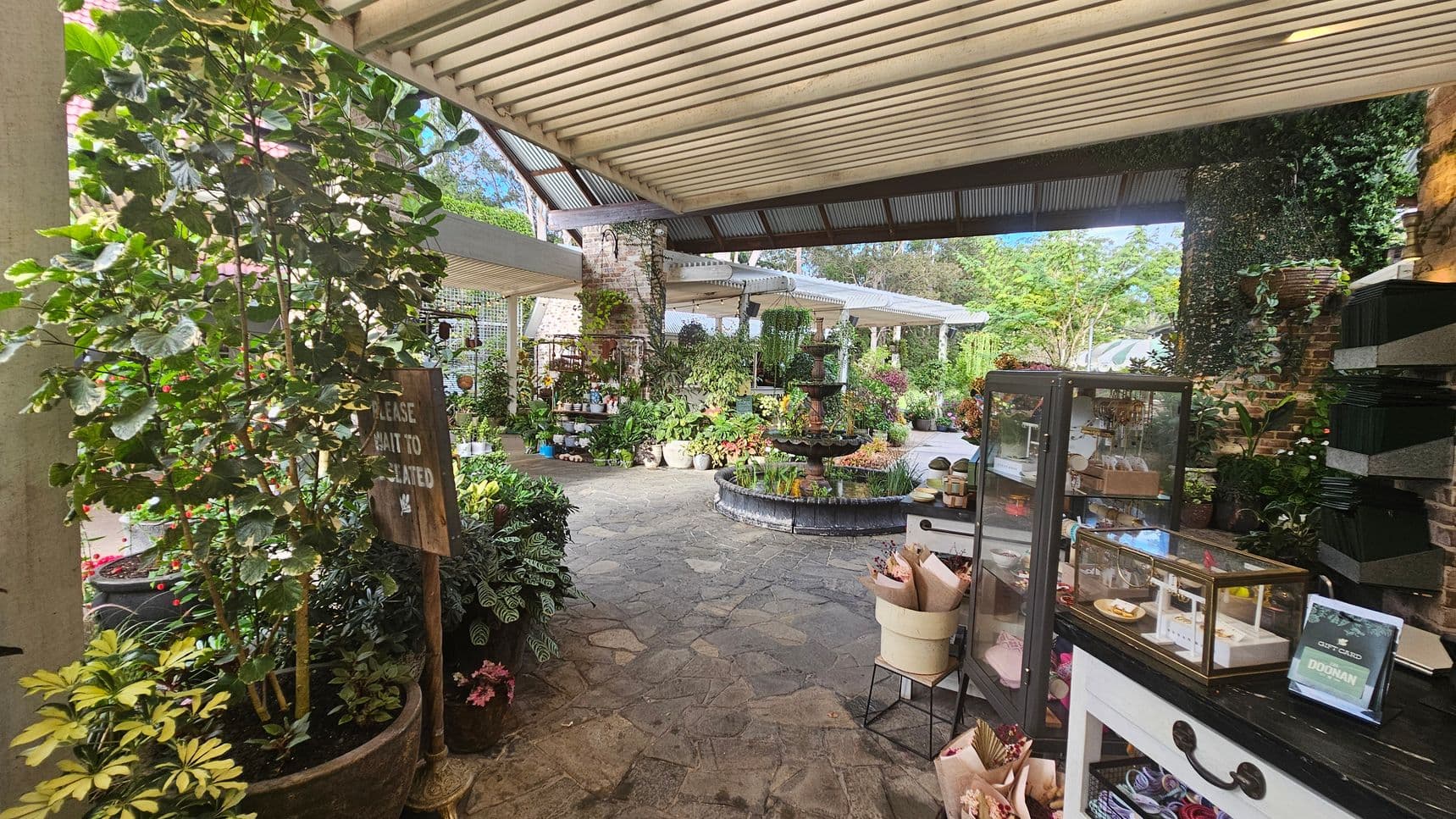 A garden centre with a variety of plants and flowers in pots on the left and centre. A sign reads "Please wait to be seated." In the middle, a stone path leads to a fountain surrounded by more plants. On the right, a display case with items for sale is on a counter, with a sign mentioning gift cards. The ceiling is covered with corrugated metal, and the floor is made of stone tiles. The background features lush greenery and a brick wall.