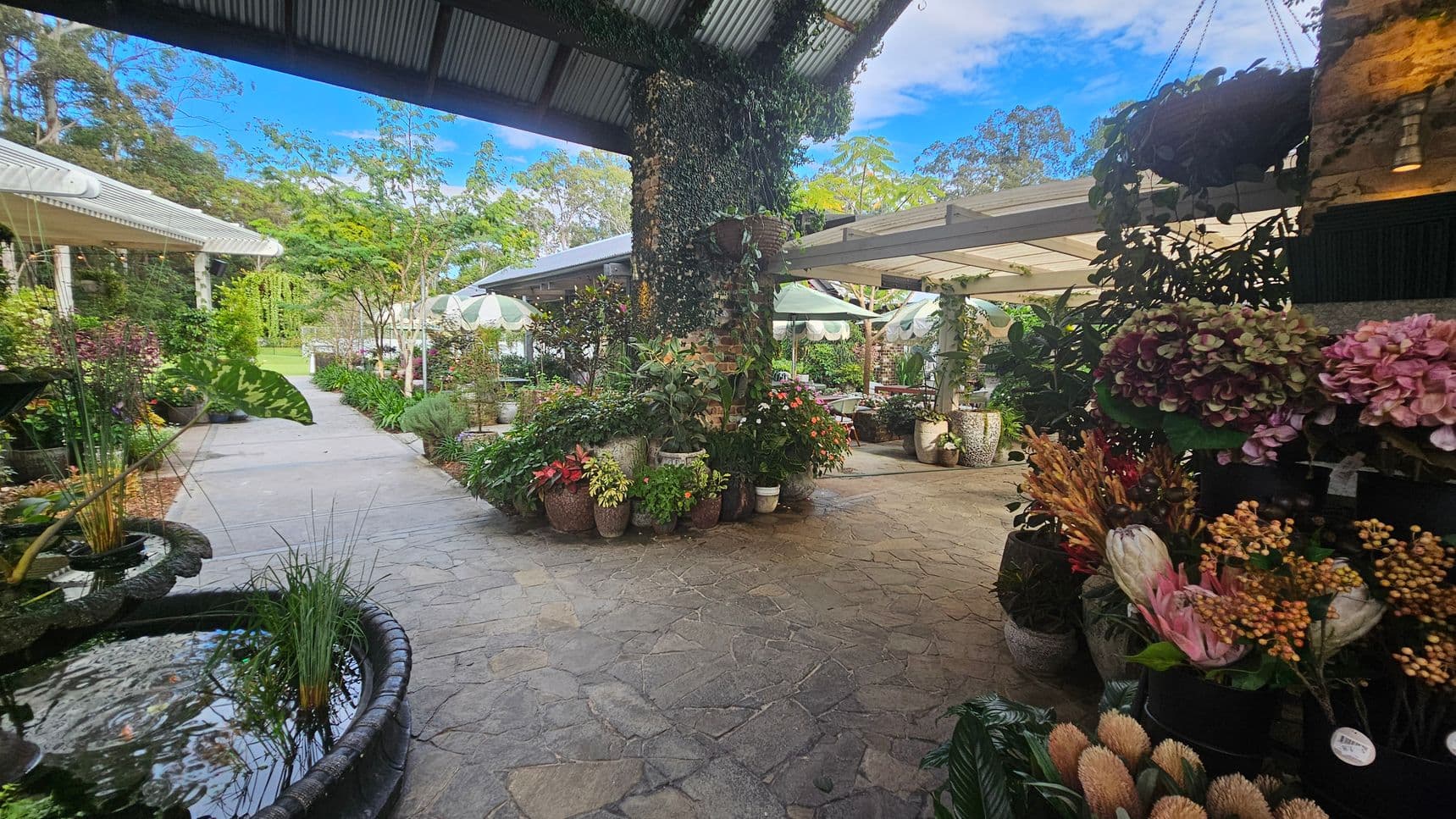 A garden centre with a variety of plants and flowers. On the left, a small pond with aquatic plants is visible. The centre features a stone pathway lined with potted plants and flowers, leading to a seating area with white umbrellas. The right side has a display of colourful flowers, including hydrangeas. The ceiling is a corrugated metal roof, and the background shows lush greenery and trees.