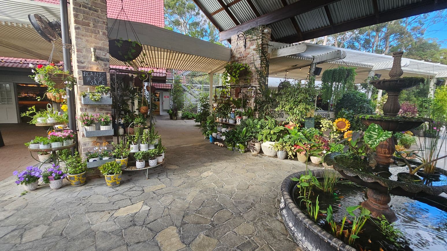 A garden centre with a variety of potted plants displayed on the left, including flowers and greenery. A brick pillar with a "Plants for Sale" sign is adorned with hanging plants. In the centre, more plants are arranged on shelves. On the right, a tiered fountain with water plants is visible. The floor is made of stone tiles, and the ceiling is corrugated metal. In the background, there are more plants and a sign for a pizzeria.