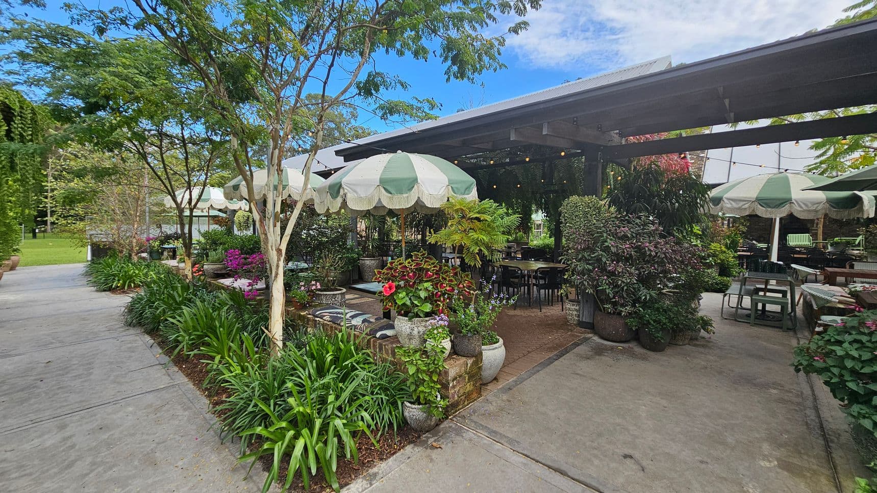 A garden café with green and white striped umbrellas shading tables and chairs. Lush greenery and colourful flowers in pots surround the seating area. A concrete path runs along the left side, bordered by plants and trees. The café structure has a metal roof, and string lights are visible under the canopy. The setting is open and inviting, with a mix of natural and man-made elements.