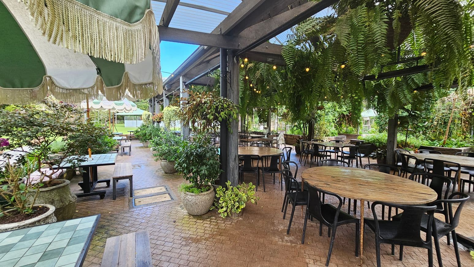 A covered outdoor seating area with round wooden tables and black chairs on the right. Hanging ferns and string lights adorn the ceiling. On the left, there are square tables with green and white checkered tops, accompanied by benches. Large potted plants are scattered throughout the space. The floor is paved with small bricks, and green and white striped awnings with fringe hang above. The background features lush greenery and more seating.