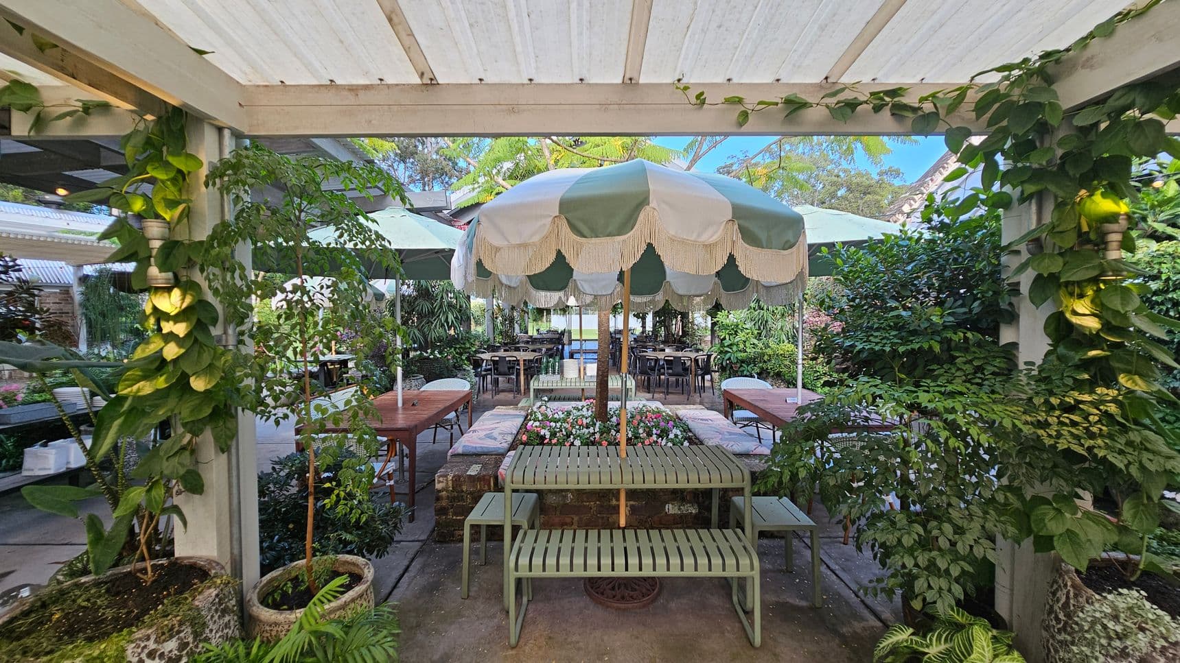 A garden setting with a central green and white striped umbrella shading a bench and table. Surrounding the area are lush green plants and foliage. On either side, there are wooden tables with chairs. The ceiling is a white pergola with climbing plants. The floor is paved, and there are planters with various plants around the space, creating a cosy, natural atmosphere.