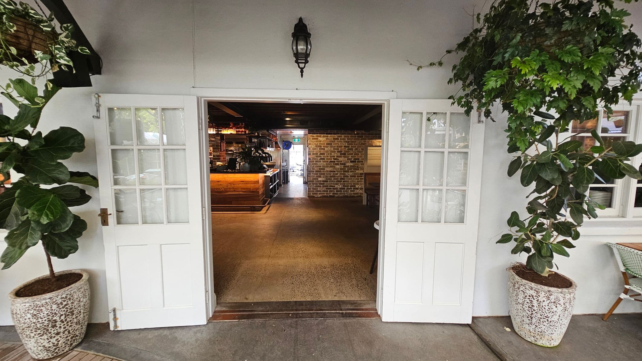 White double doors with glass panes open into an interior space. On either side of the entrance, there are large potted plants with broad green leaves. Inside, a wooden counter is visible to the left, and a brick wall is at the back. The floor is a polished concrete, and a black lantern-style light fixture is mounted above the doors. A small table and chair are partially visible on the right.