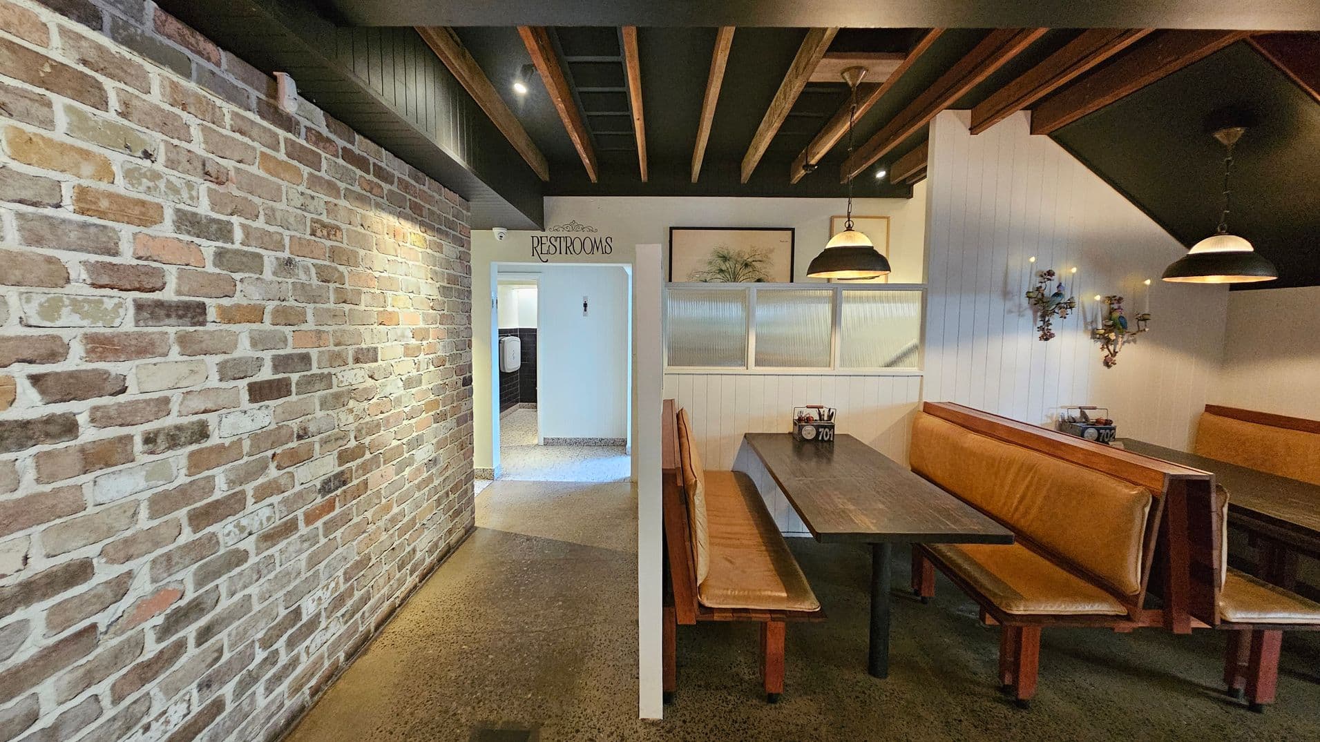 A restaurant interior with a rustic brick wall on the left. In the centre, an entrance marked "Restrooms" leads to a hallway. On the right, there are wooden booths with brown leather seating and dark tables. Above the booths, pendant lights hang from a ceiling with exposed wooden beams. The wall behind the booths is white with decorative sconces. The floor is polished concrete.
