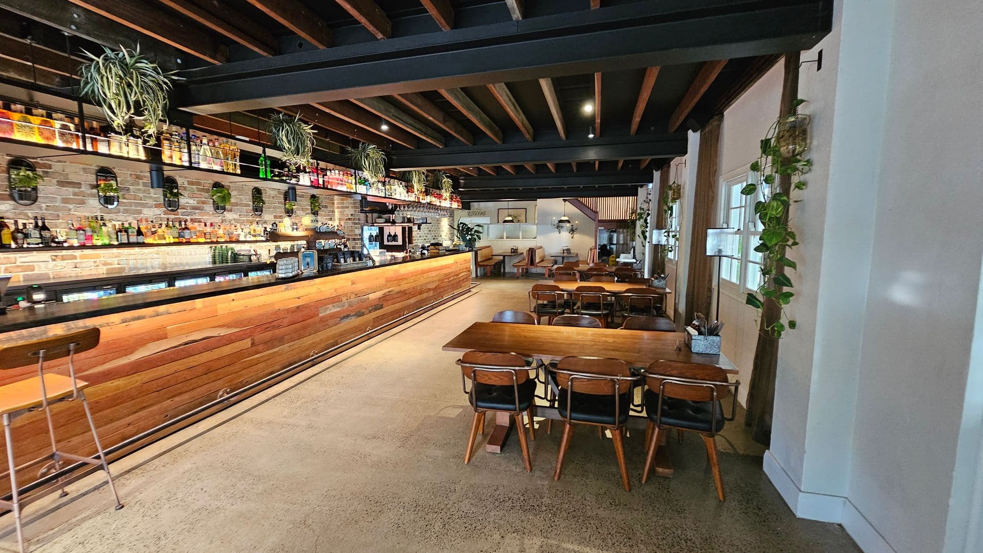 A spacious bar interior with a long wooden counter on the left, lined with various bottles and plants above. The back wall is exposed brick with additional plants and mirrors. On the right, there are wooden tables and chairs, with more plants hanging on the walls. The ceiling features exposed wooden beams, and the floor is polished concrete. In the background, there are more seating areas with booths and pendant lights.