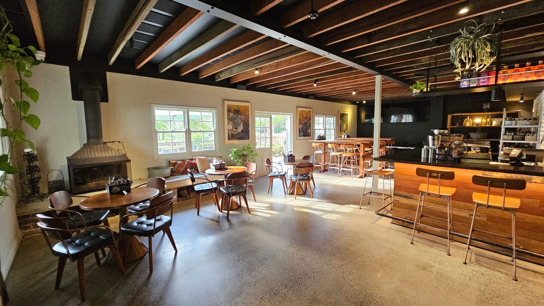A cosy café interior with a polished concrete floor and a ceiling featuring exposed wooden beams. On the left, there's a fireplace next to a round wooden table with chairs. Large windows allow natural light to illuminate the space. In the centre, more tables and chairs are arranged, with a plant adding greenery. On the right, a wooden bar with stools is visible, with shelves and a coffee machine behind it. A hanging plant is suspended from the ceiling.