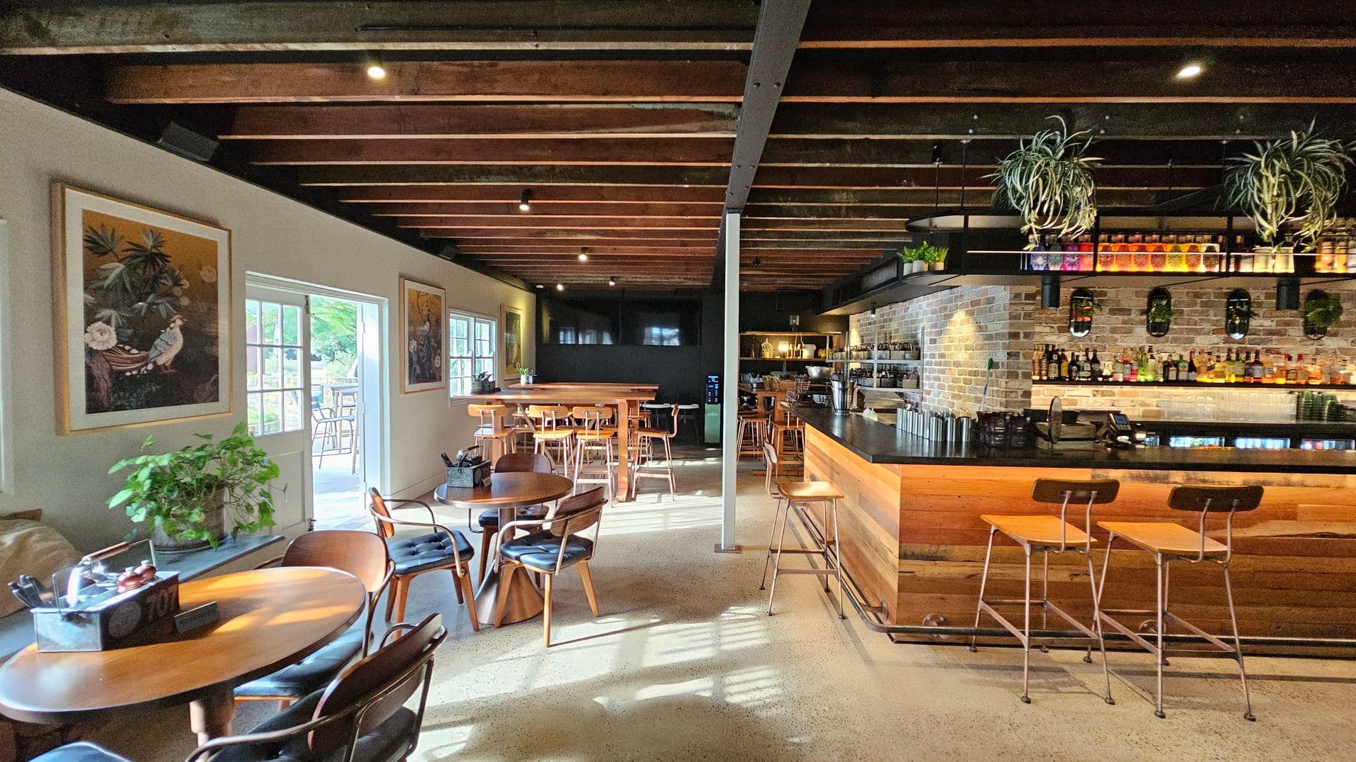 A cosy café interior with wooden beams on the ceiling. On the left, there are round wooden tables with chairs, a plant, and framed artwork on the wall. Sunlight streams through a door leading outside. In the middle, more tables and chairs are arranged. On the right, a bar with a brick wall backdrop displays various bottles, with plants hanging above. The floor is polished concrete, and the atmosphere is warm and inviting.
