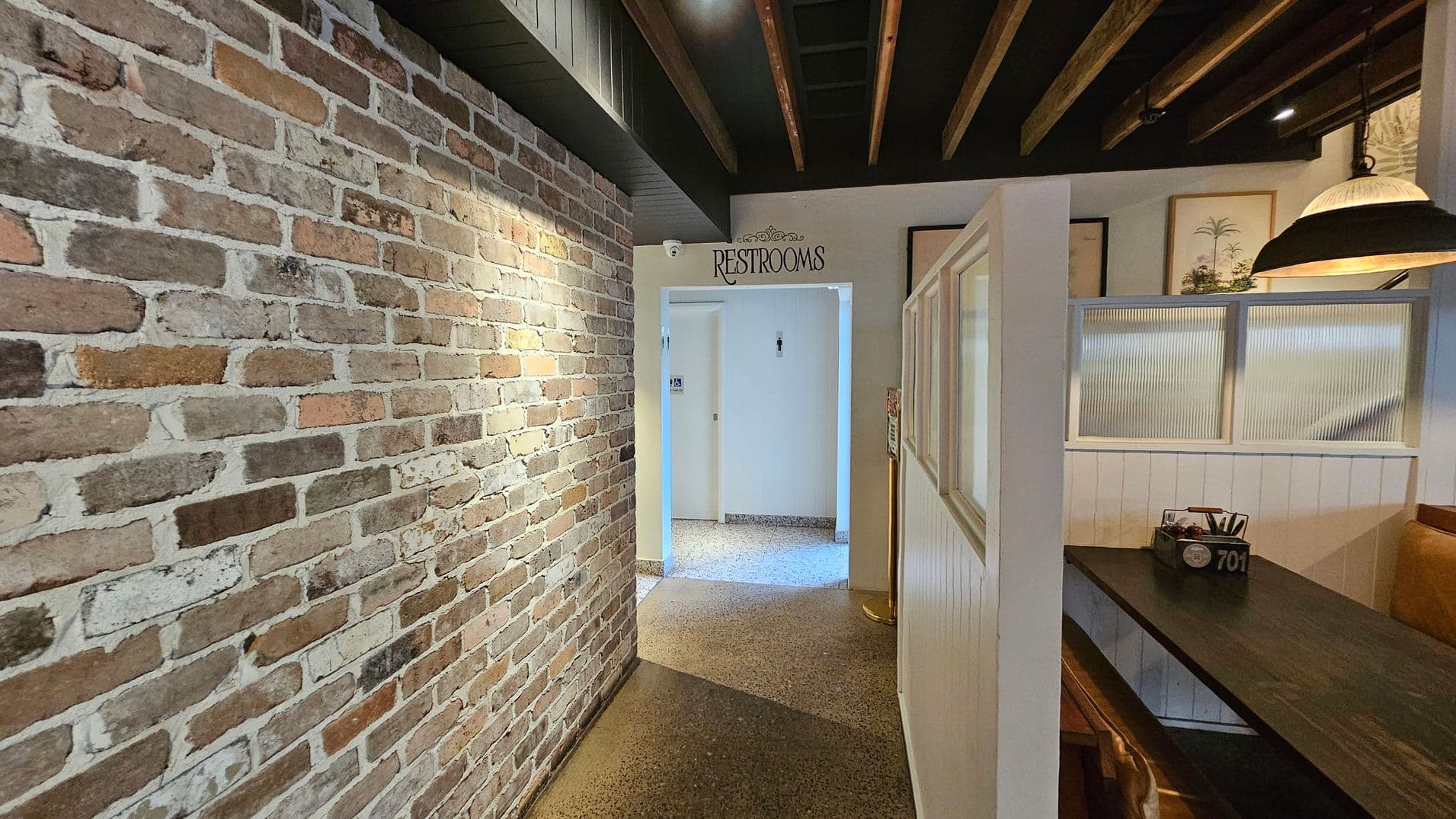 A hallway with a rustic brick wall on the left and a polished stone floor. On the right, there is a dining booth with a dark wooden table and a partition with frosted glass panels. Above the booth, a large pendant light hangs from the ceiling, which features exposed wooden beams. In the centre, a doorway leads to restrooms, marked by a sign above. The walls near the restrooms are painted white, and there are framed pictures on the wall near the booth.
