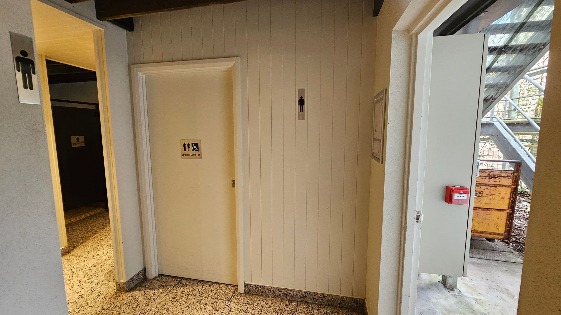 A corridor with a tiled floor and a white panelled wall. On the left, there is a door with a male restroom sign. In the centre, another door has a sign indicating a unisex and accessible toilet. On the right, an open door leads outside, revealing a metal staircase and a rusty container. A red fire alarm is mounted on the wall near the open door. The ceiling has exposed beams.