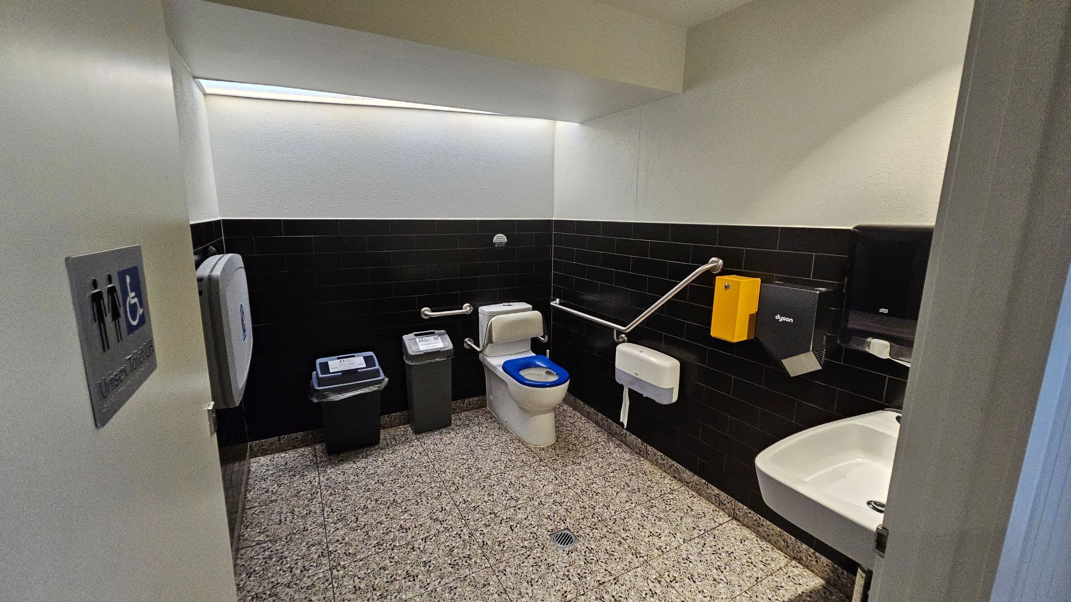 A public accessible toilet with a white toilet featuring a blue seat cover in the centre. To the left, there is a bin and a baby changing station. The walls are tiled with black tiles halfway up, and the floor is speckled grey. On the right, a white sink is mounted on the wall with a hand dryer and paper towel dispenser above it. Stainless steel grab bars are installed around the toilet. The ceiling is plain white, and a sign on the door indicates it is a unisex toilet.