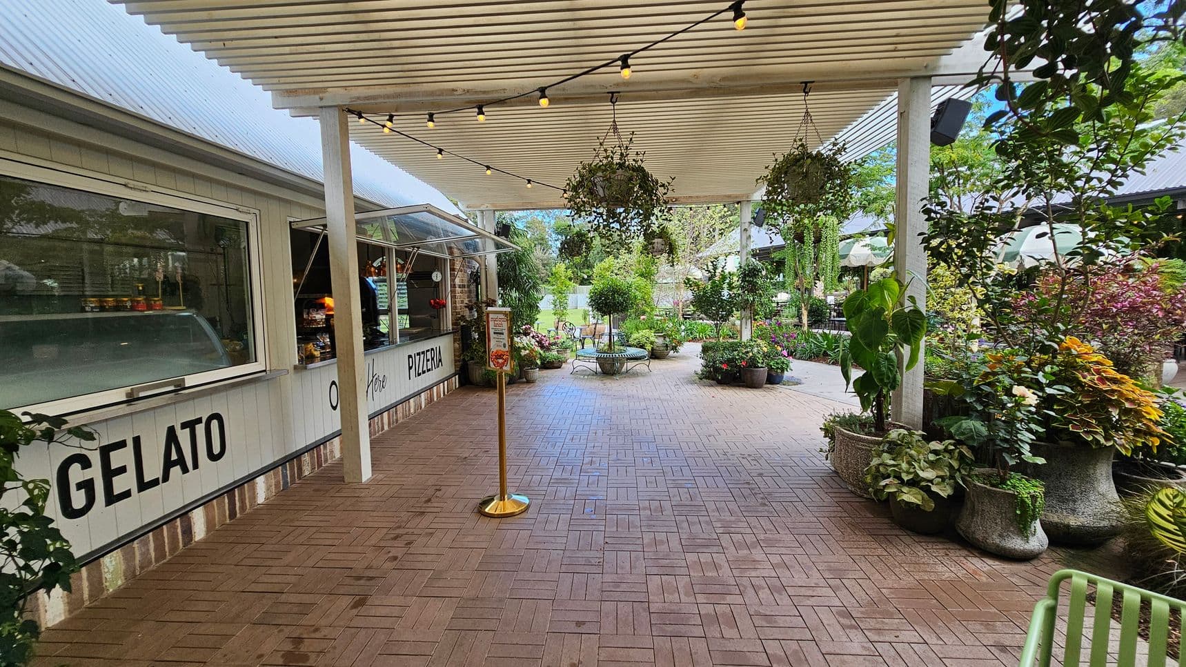A covered outdoor area with a gelato and pizzeria counter on the left, featuring a white facade and large windows. The ceiling has a slatted design with string lights and hanging plants. The floor is paved with interlocking bricks. On the right, there are various potted plants and greenery. In the background, more plants and a seating area with a bench are visible, creating a garden-like atmosphere.