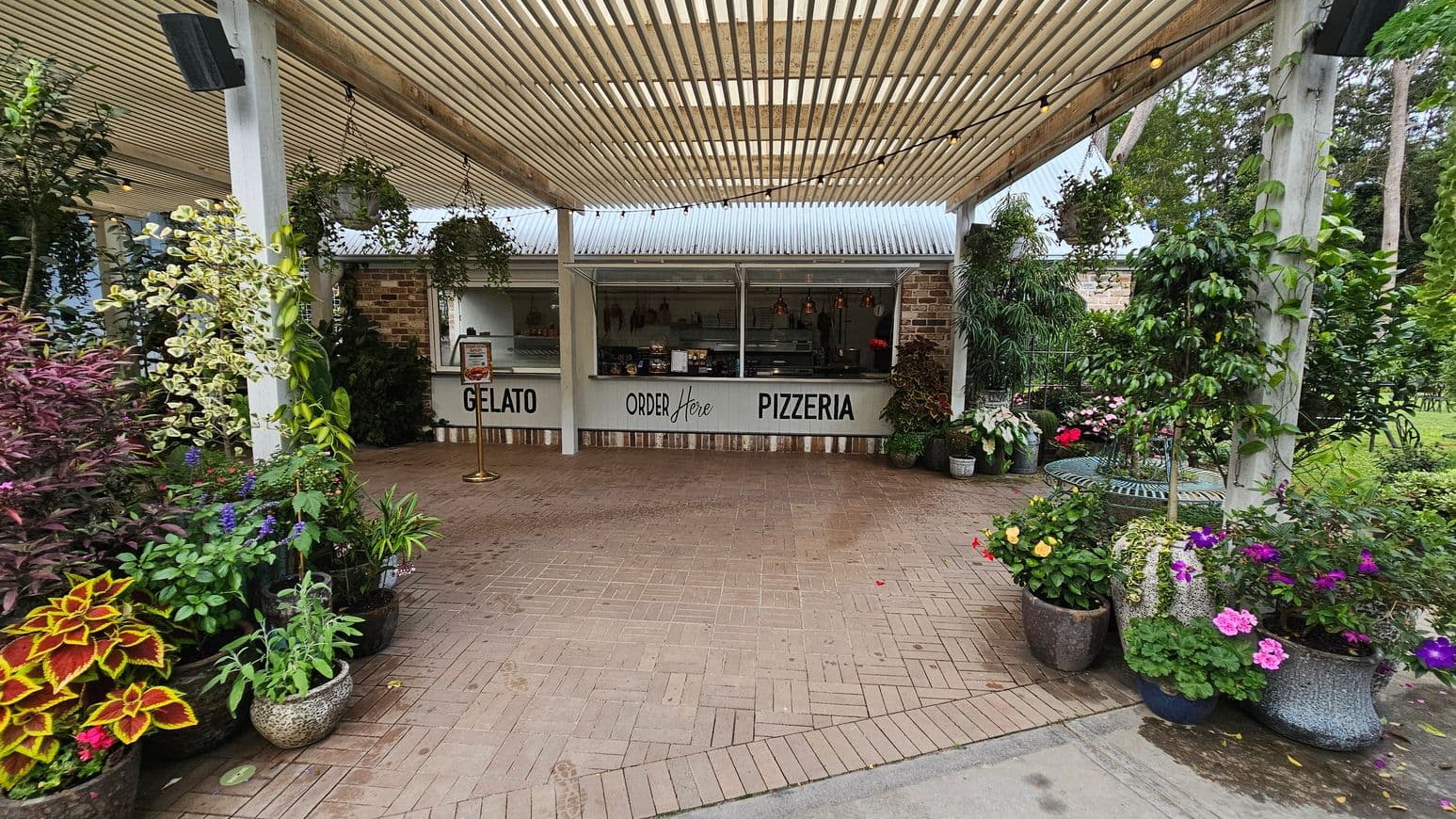 A pizzeria and gelato stand with a sign reading "Order Here" is in the centre, featuring a brick wall and a corrugated metal roof. The foreground has a paved area with a variety of potted plants and flowers on both sides, including colourful blooms and lush greenery. The ceiling is a slatted pergola with string lights. In the background, there are more plants and trees, creating a garden-like atmosphere.