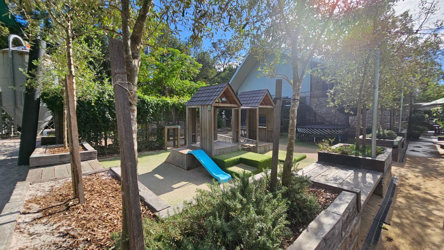 A children's play area with a wooden structure featuring two small peaked roofs and a blue slide in the centre. The ground is covered with sand and bordered by low stone walls. Trees and shrubs are planted around the area, providing shade and greenery. In the background, there is a large building with a corrugated metal exterior. To the left, a tall metal structure is partially visible. The area is well-maintained and designed for outdoor play.