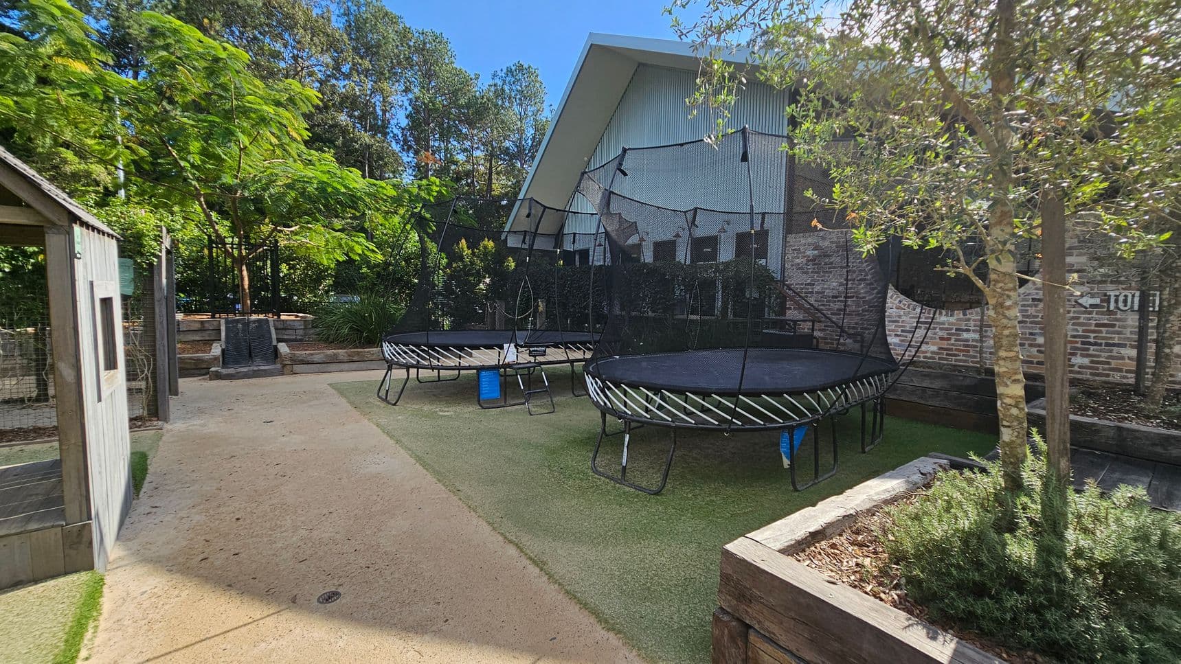 A garden area with two trampolines in the centre, each surrounded by safety nets. On the left, there's a small wooden structure resembling a shed. The ground is a mix of concrete and artificial grass. In the foreground, a planter box with a small tree and shrubs. The background features a large building with a slanted roof and brick walls, surrounded by tall trees and greenery.