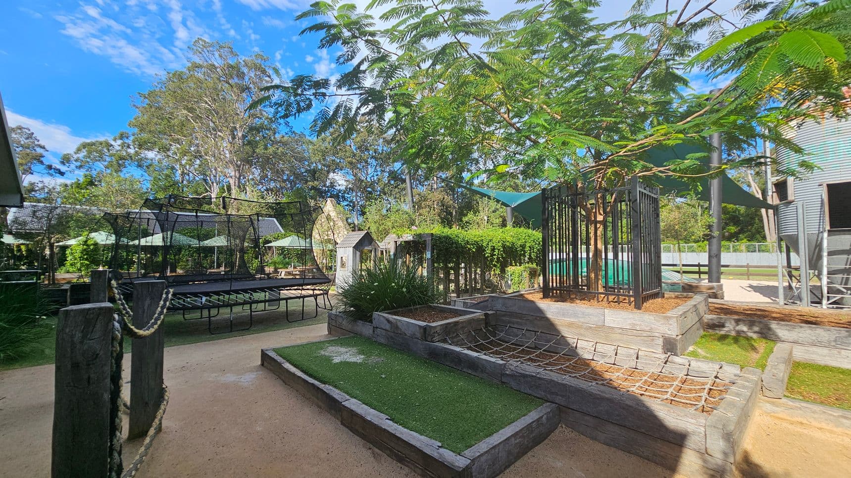 Outdoor play area with various structures. On the left, a large trampoline with a netted enclosure is surrounded by wooden posts connected by ropes. In the centre, a small wooden playhouse and a garden with lush greenery. On the right, a tree is enclosed by a metal fence, and a climbing net is attached to raised wooden platforms. The ground is a mix of sand and artificial grass. In the background, tall trees and green shade sails provide cover.