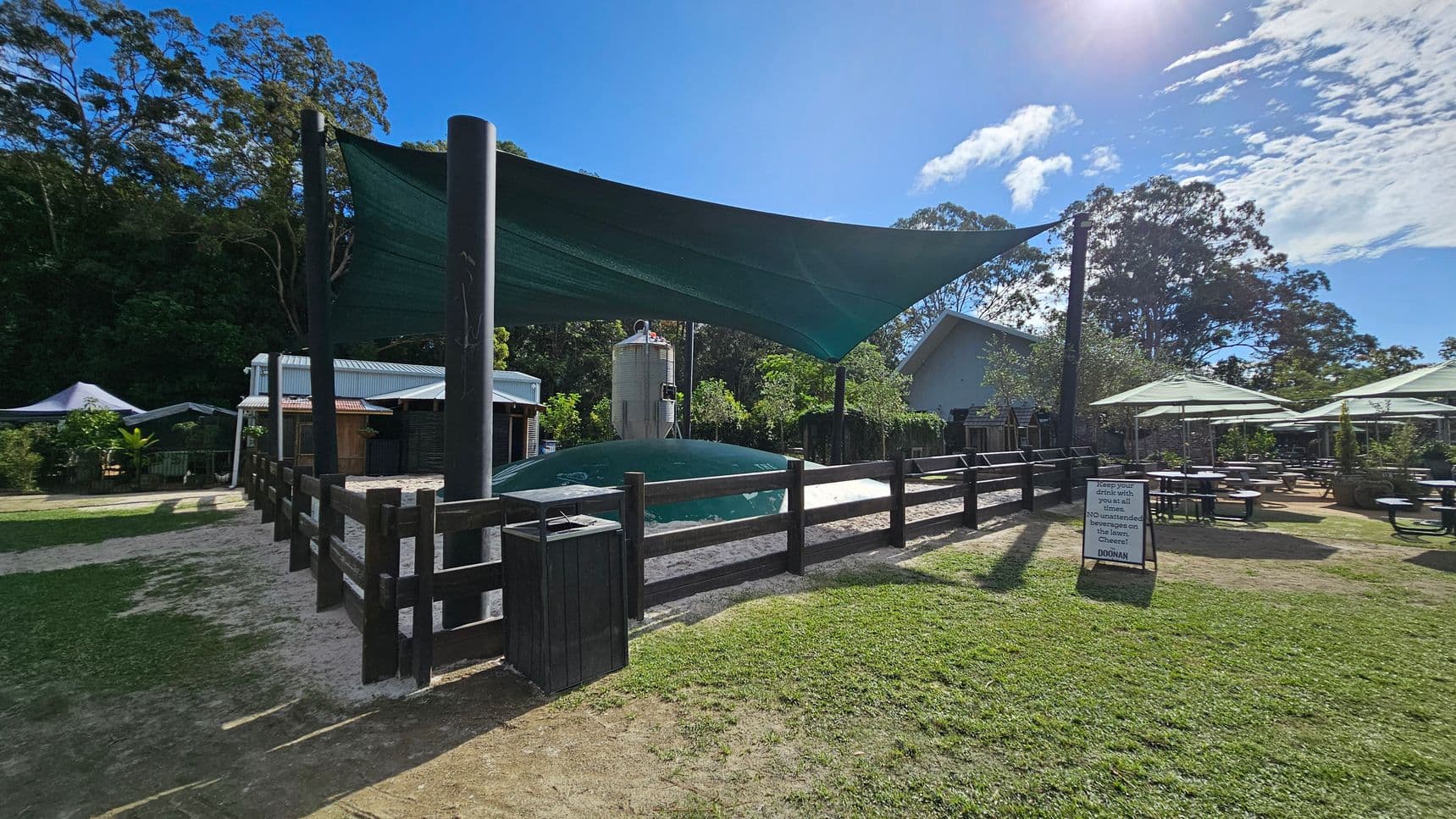 A fenced area with a large, inflated play structure covered by a green shade sail is in the centre. To the left, there are small buildings with metal roofs surrounded by trees. On the right, outdoor seating with tables and umbrellas is visible on a grassy area. A sign with text is placed near the fence. The background features tall trees and a clear sky. The ground is a mix of grass and dirt.