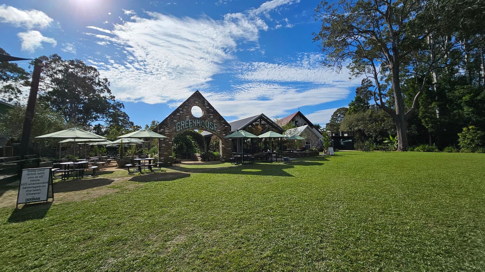 A grassy area with a building in the centre featuring a triangular roof and a circular window, labelled "GREENHOUSE". Several outdoor tables with umbrellas are on the left and right. A sign on the left reads, "Keep your drink with you at all times. NO unattended beverages on the lawn. Cheers! DOONAN". Tall trees surround the area, providing shade and greenery.