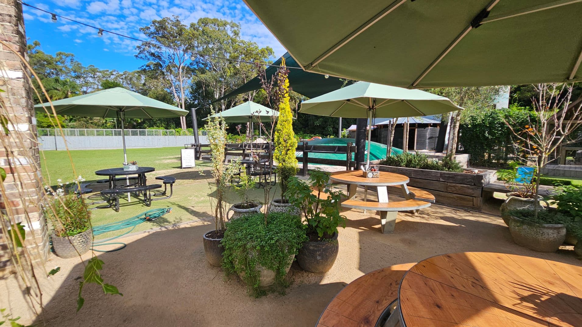 Outdoor seating area with round wooden tables and green umbrellas. In the foreground, several potted plants are arranged on a sandy surface. To the left, a brick wall is partially visible. In the background, a grassy area with more tables and a fence is surrounded by tall trees. A garden hose is coiled on the ground near a black metal table. Overhead, string lights are strung across the area.