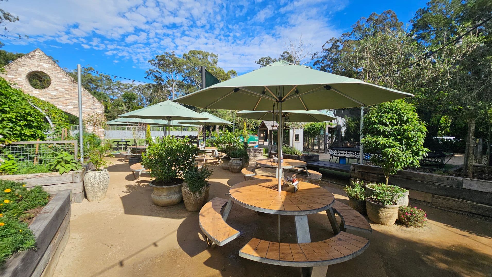 Outdoor seating area with round wooden tables and green umbrellas in the centre. Potted plants and greenery surround the space. On the left, a brick structure with a circular window is partially covered in vines. In the background, trees and a wooden fence are visible. The ground is sandy, and string lights are strung overhead.