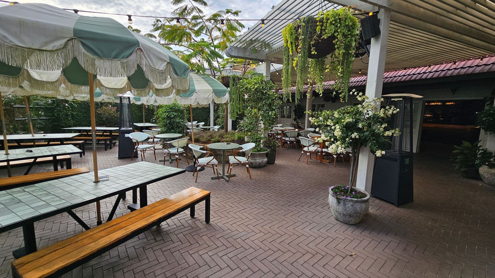 Outdoor seating area with a herringbone-patterned brick floor. On the left, there are long tables with wooden benches under large green and white striped umbrellas. In the centre, round tables with wicker chairs are surrounded by potted plants. On the right, a pergola with hanging plants provides shade. A tall outdoor heater stands nearby. The background features lush greenery and a building with a red-tiled roof.