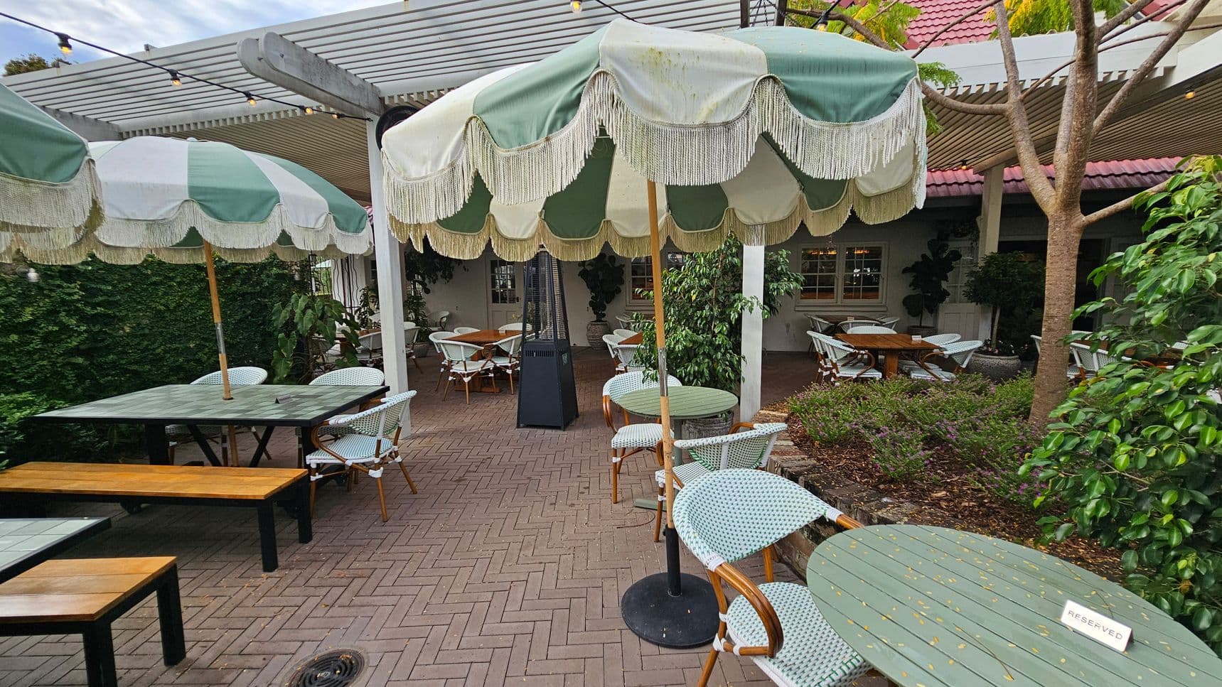 Outdoor café setting with green and white striped umbrellas featuring fringe, covering round tables with wicker chairs. A "Reserved" sign is on one table. Wooden benches accompany rectangular tables on the left. The ground is paved with herringbone-patterned bricks. A pergola with string lights is overhead. Lush greenery and potted plants surround the area, with a tree on the right. A building with a red-tiled roof and windows is in the background.