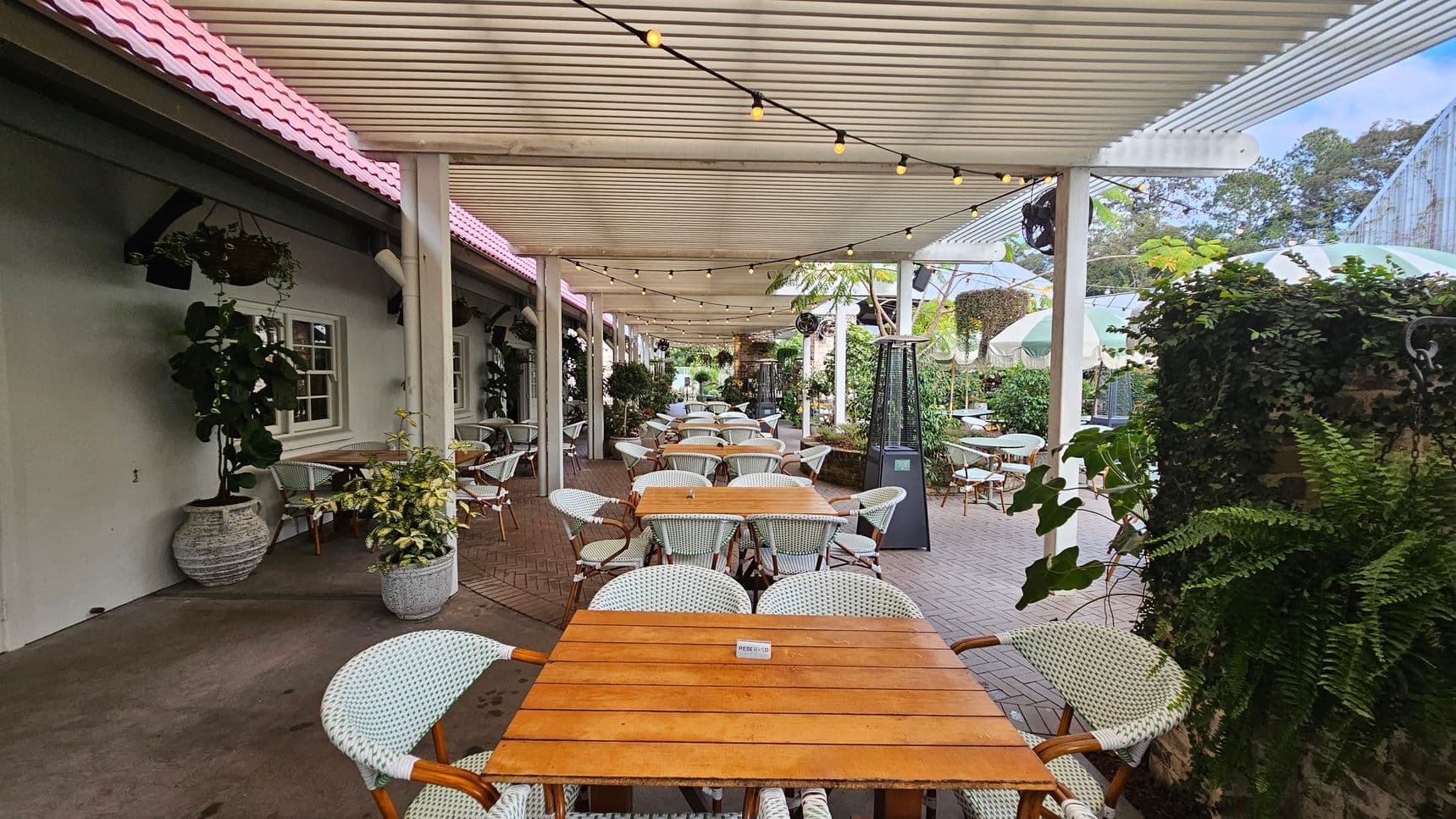 Outdoor dining area with wooden tables and white wicker chairs arranged in rows. A pergola with string lights covers the space. On the left, a building with a pink-tiled roof and green walls features windows and hanging plants. On the right, green foliage and potted plants line the area. The floor is a mix of concrete and brick, and patio heaters are visible. In the background, more tables and greenery are present, creating a lush atmosphere.