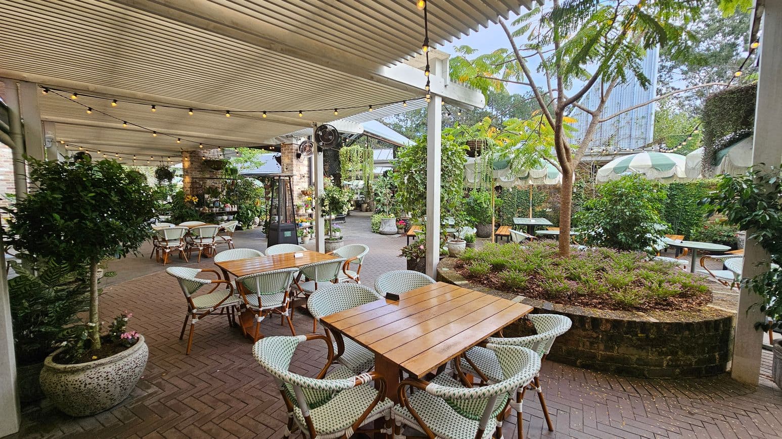 An outdoor dining area with wooden tables and wicker chairs on a herringbone brick floor. The left side features a covered patio with string lights and potted plants. In the centre, a round brick planter with a small tree and shrubs. The right side has more tables under green and white striped umbrellas. The background includes lush greenery and a tall building partially visible. The ceiling is a slatted pergola.
