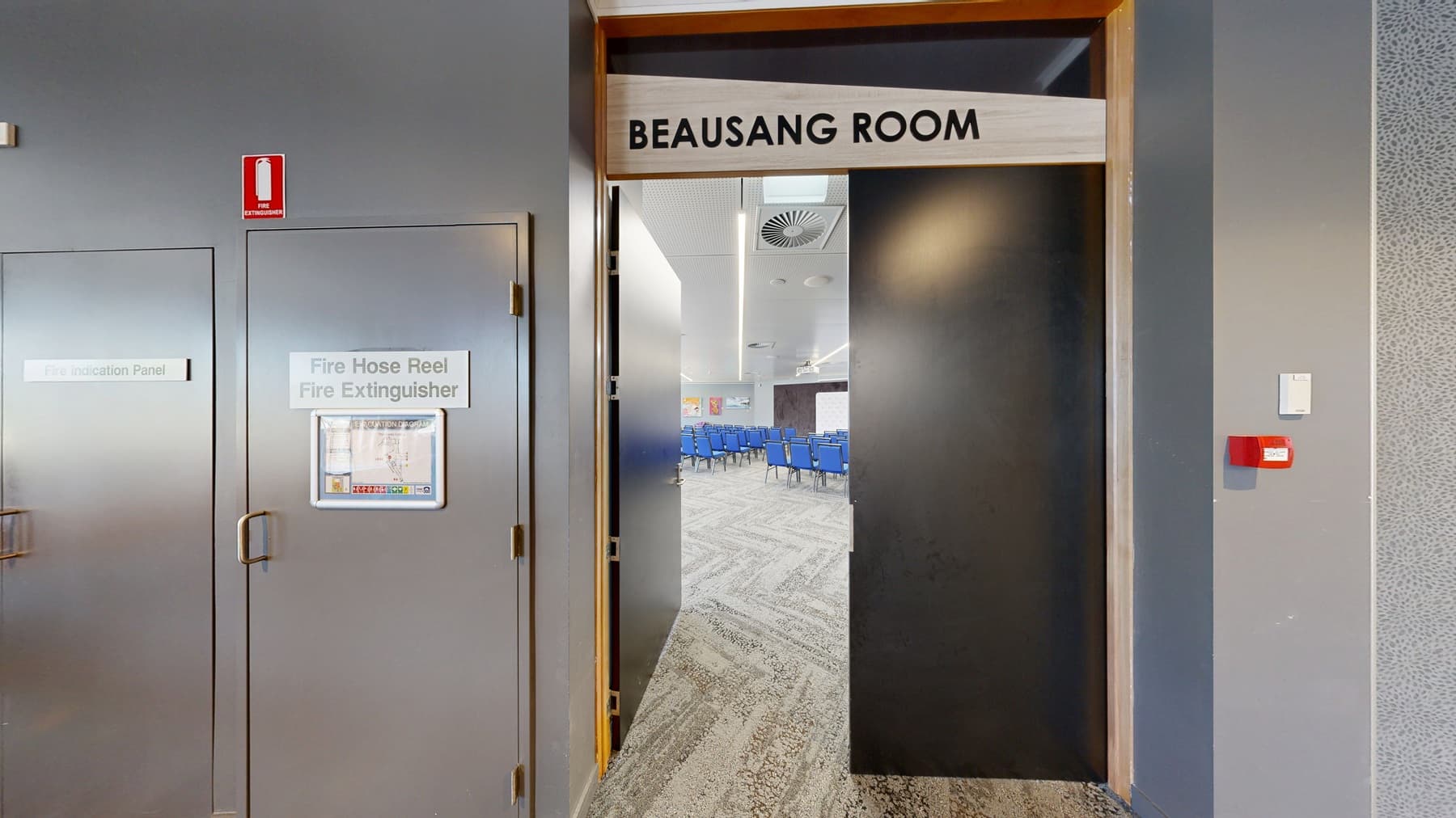 On the left, a room with rows of blue chairs facing forward, with colourful artwork on the walls. The floor is carpeted in a light grey pattern. On the right, a hallway with a brown and green patterned carpet, featuring several large potted plants. A digital screen is mounted on the wall displaying an advertisement. The ceiling has recessed lighting and air vents. A partially open door with a sign above it reads "ROOM".