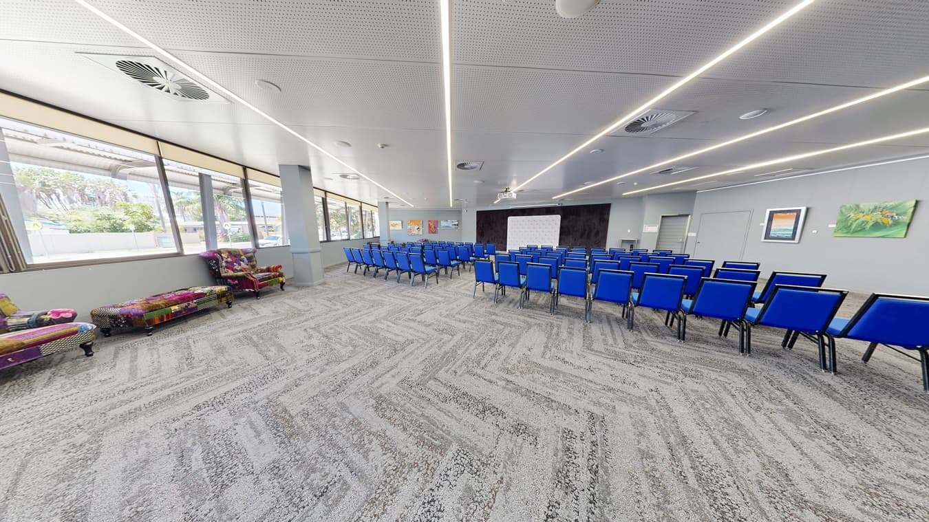 A spacious conference room with a grey patterned carpet and a white ceiling featuring linear lights. On the right, rows of blue chairs are arranged facing a white screen at the front. The left side has colourful, patterned sofas near large windows that let in natural light. The walls are adorned with framed artwork.