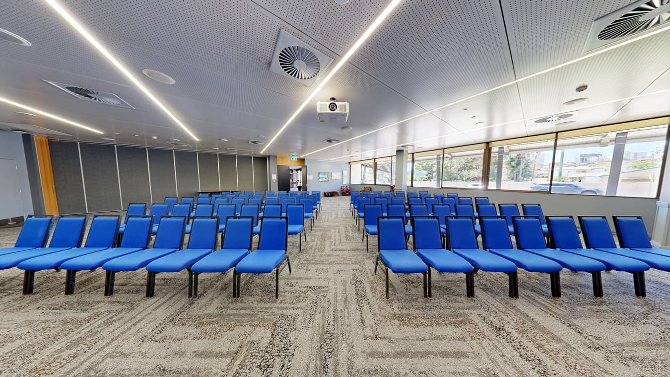 Rows of blue chairs are arranged in a conference room. On the left, there are grey partition walls. On the right, large windows allow natural light to enter, showing a view of trees and buildings outside. The ceiling is white with recessed lighting and air vents. The floor is carpeted in a patterned design. At the back, there is a projector mounted on the ceiling and an exit sign above a door.