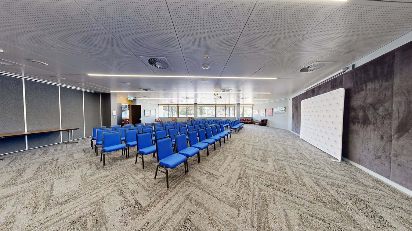 A conference room with rows of blue chairs arranged in the centre. On the left, there is a long table against a wall with vertical panels. The right side features a large white display board. The background has large windows allowing natural light, and the floor is covered with a patterned carpet. The ceiling is white with recessed lighting and circular air vents.