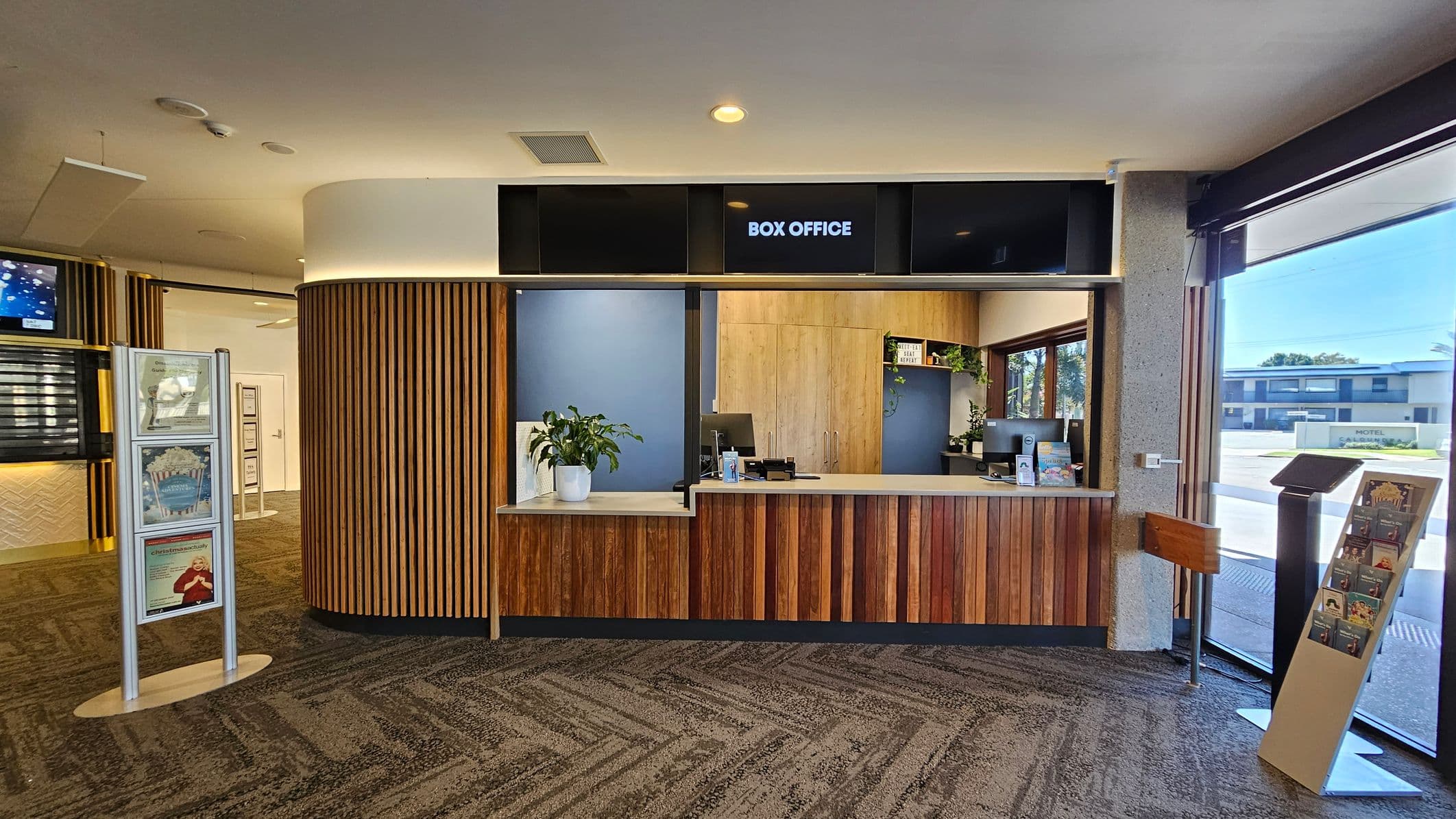 Entrance to a building with large glass doors. On the left, a reflection shows palm trees and a street. The right side features wooden panelling with part of the word "Box" visible. Inside, a carpeted area with a circular light fixture on the ceiling, a reception desk, and digital screens. A black mat at the entrance reads "the events centre CALOUNDRA | SUNSHINE COAST." The floor outside is light-coloured with a textured surface.